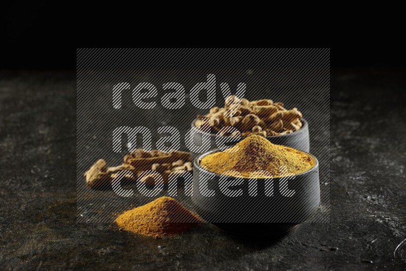 2 pottery black bowls, one full of turmeric powder and the other full of dried turmeric whole fingers on a textured black flooring