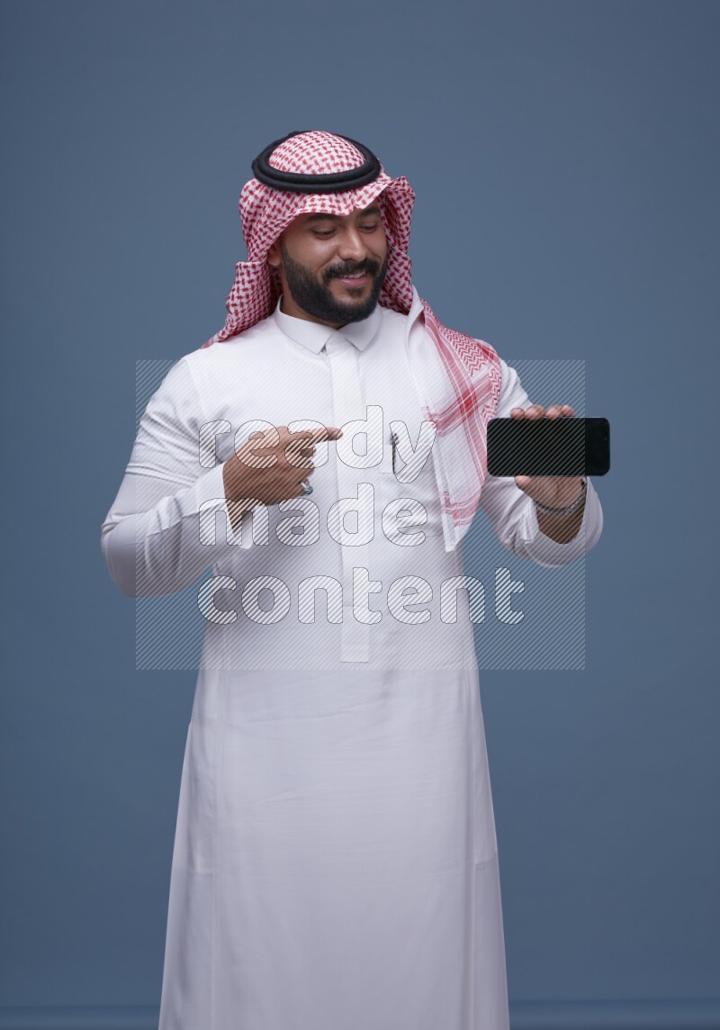 A man Showing his smartphone Screen on Blue Background wearing Saudi Thob