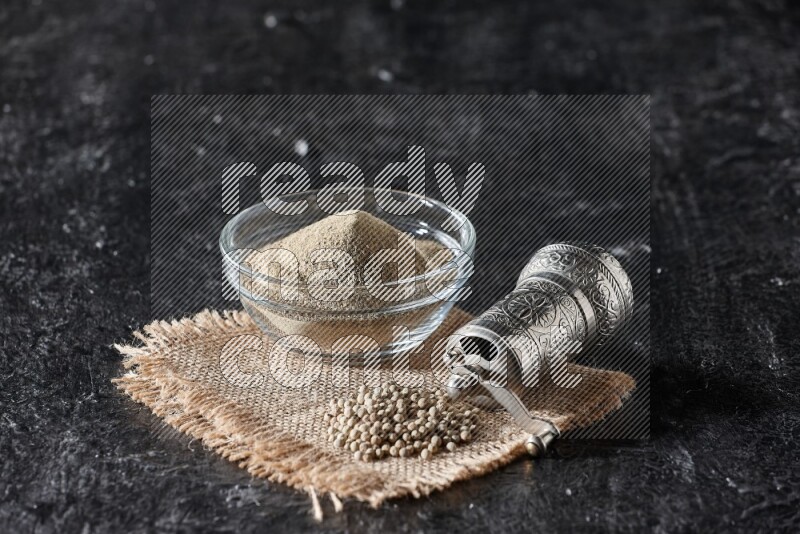 A glass bowl full of white pepper powder with white pepper beads on a burlap piece of fabric and a metal grinder on textured black flooring
