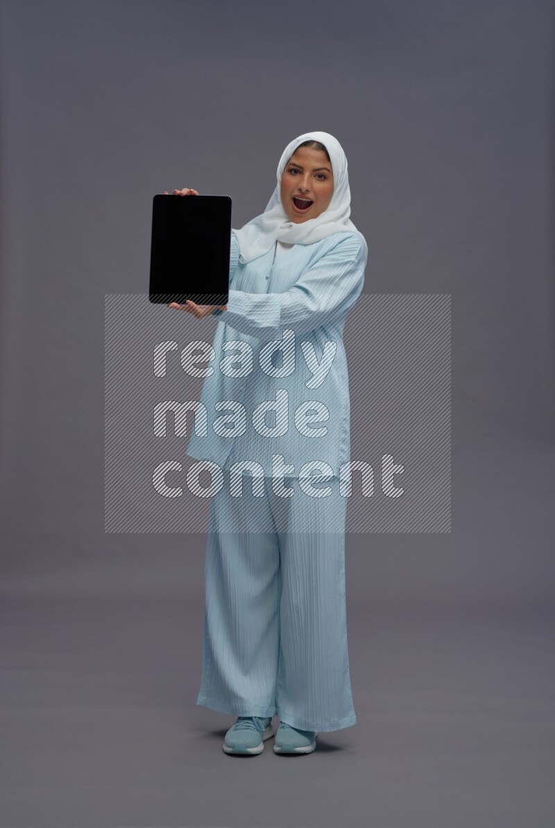 Saudi woman wearing hijab clothes standing showing tablet to camera on gray background