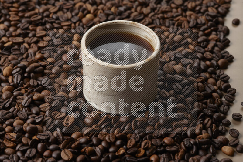 A beige pottery cup of coffee surrounded by roasted coffee beans on beige background