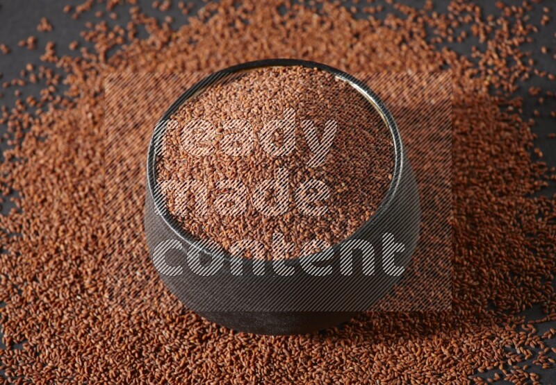 A black pottery bowl full of garden cress seeds surrounded by seeds on a black flooring