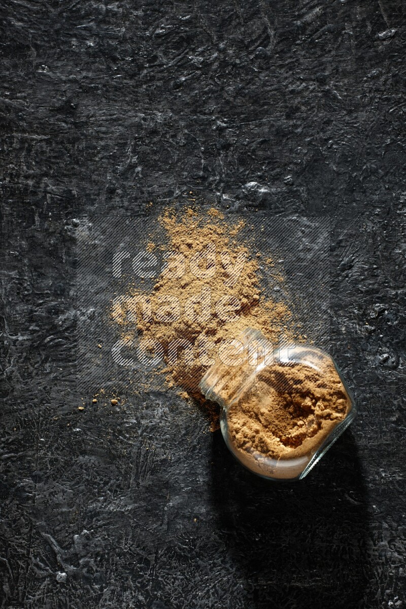 A flipped glass spice jar full of allspice powder and powder spilled out of it on a textured black flooring