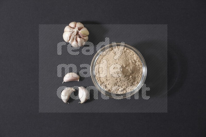 A glass bowl full of garlic powder with garlic bulb and some cloves beside it on a black flooring