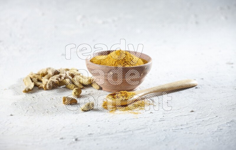A wooden bowl and wooden spoon full of turmeric powder with dried turmeric fingers beside it on textured white flooring