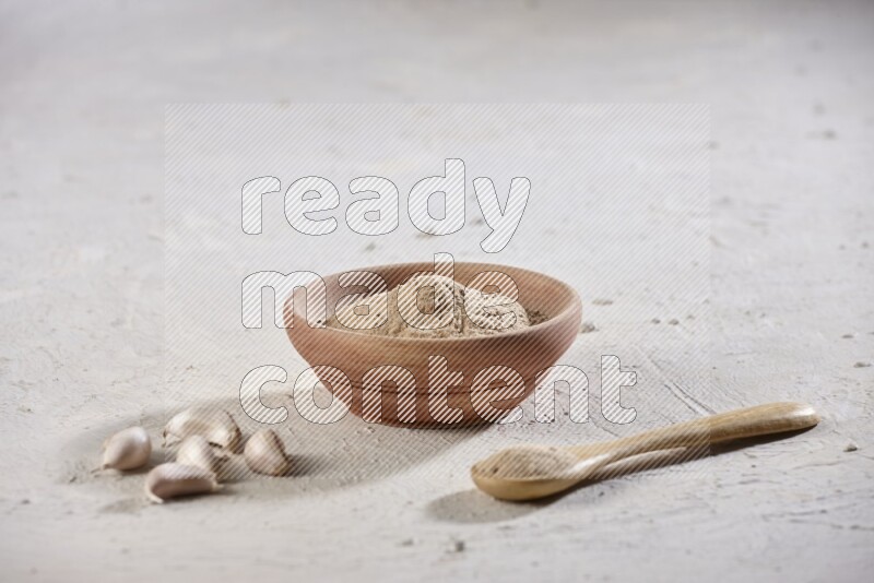 A wooden bowl and spoon full of garlic powder and beside it garlic cloves on a textured white flooring