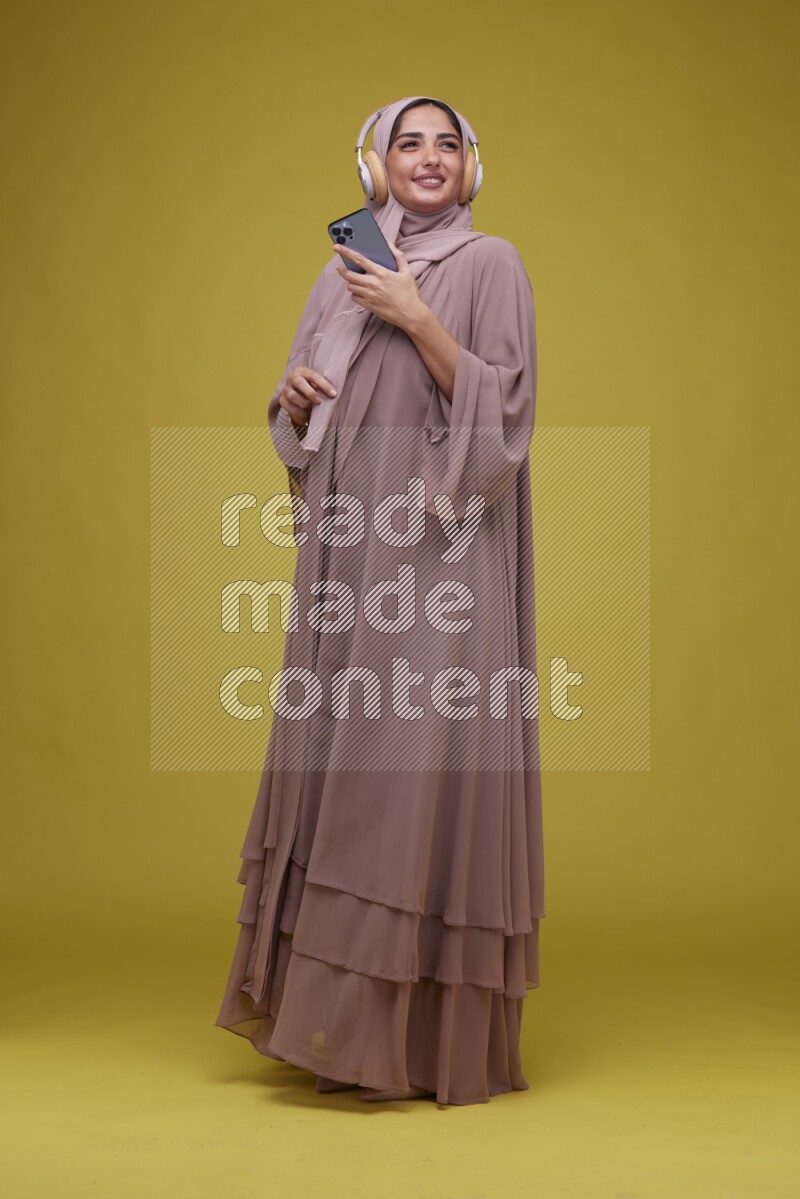 A woman Listening to Music on a Yellow Background wearing Brown Abaya with Hijab