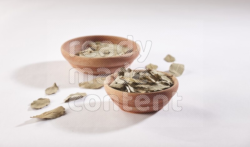 2 wooden bowls full of dried bay leaves with more leaves spread on white flooring