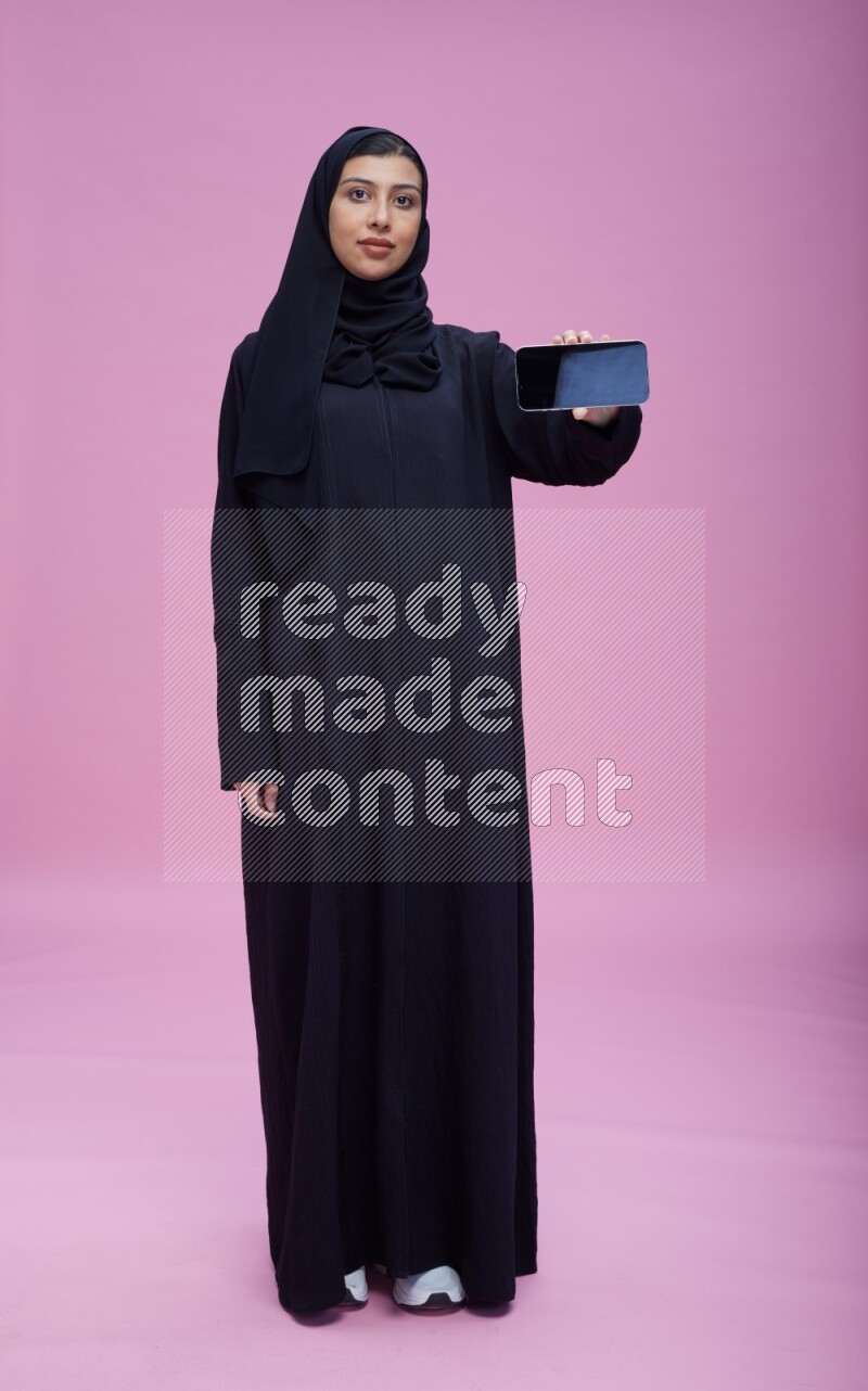 Saudi woman wearing Abaya standing showing phone to camera on pink background