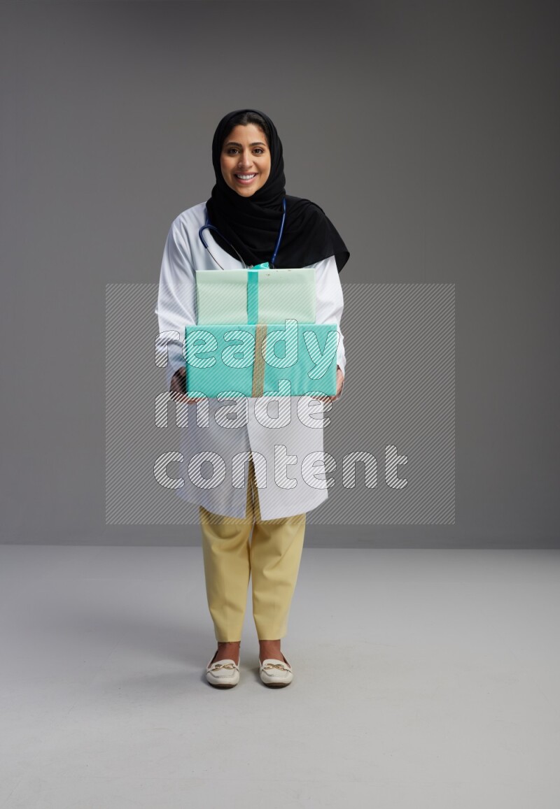 Saudi woman wearing lab coat with stethoscope standing holding gift box on Gray background