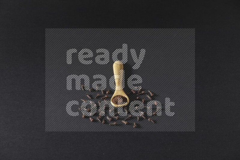 A wooden spoon full of cloves powder with spreaded whole cloves on a black flooring