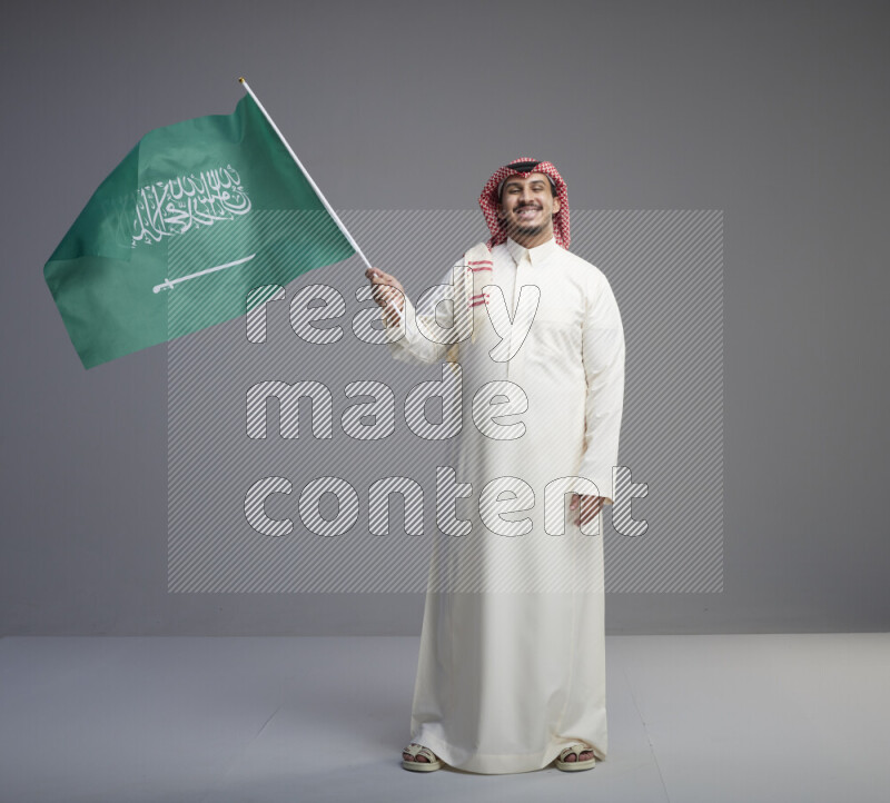 A Saudi man standing wearing thob and red shomag raising big Saudi flag on gray background