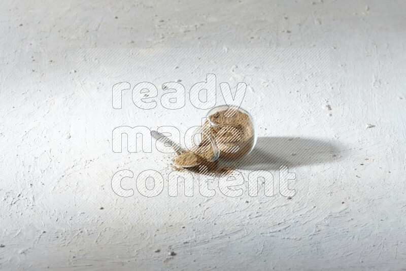 A flipped glass spice jar and a metal spoon full of cumin powder and powder spilled out on textured white flooring