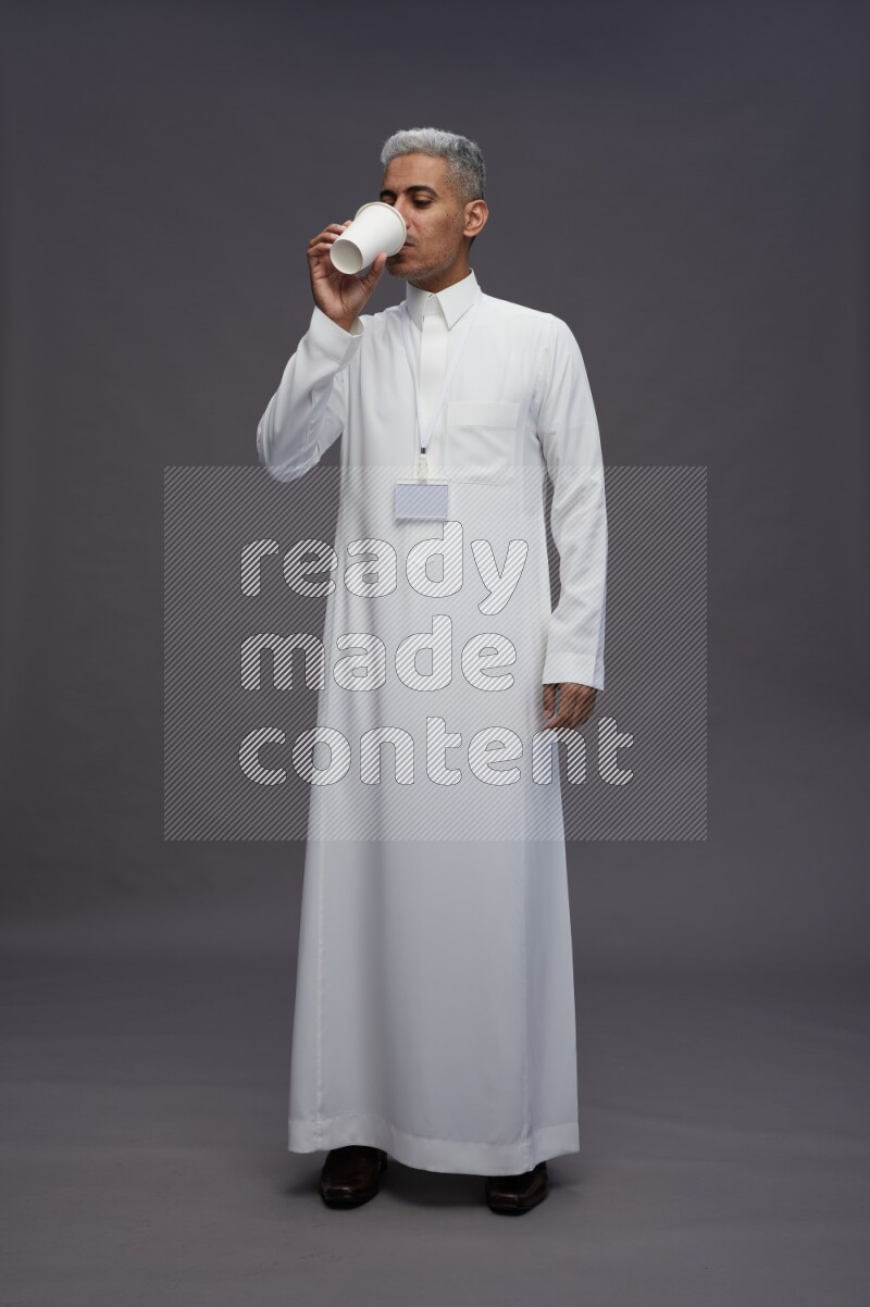 Saudi man wearing thob with neck strap employee badge standing holding paper cup on gray background