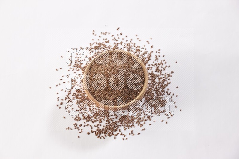A wooden bowl full of flax seeds surrounded by flax seeds on a white flooring