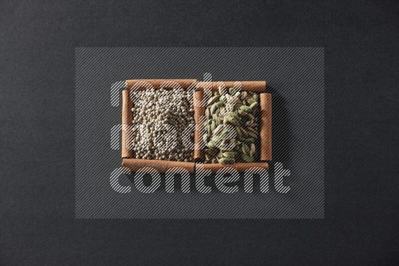 مربعان من أعواد القرفة ممتلئان بحبوب الفلفل الأبيض وحبوب الهيل علي خلفية سوداء