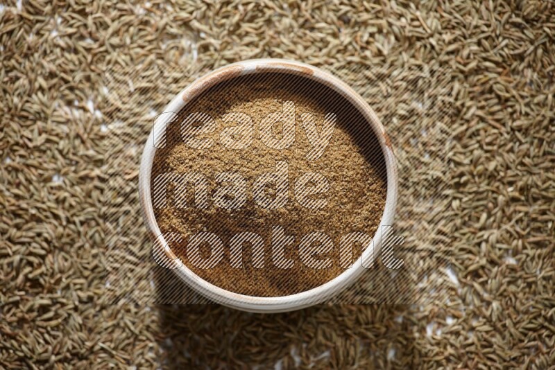 وعاء بيج ممتلئ ببودرة الكمون مع حبوب الكمون تحت الوعاء على خلفية بيضاء