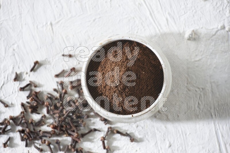 A beige ceramic bowl full of cloves powder and whole cloves on a white flooring