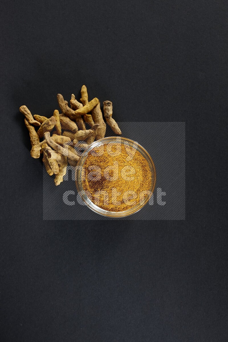 A glass bowl full of turmeric powder and dried whole fingers beside it on black flooring