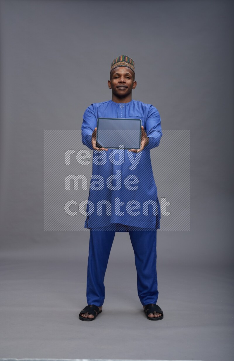 Man wearing Nigerian outfit standing showing tablet to camera on gray background