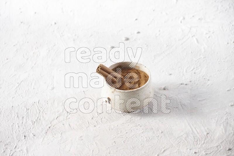 Ceramic beige bowl full of cinnamon powder with a cinnamon stick on a textured white background