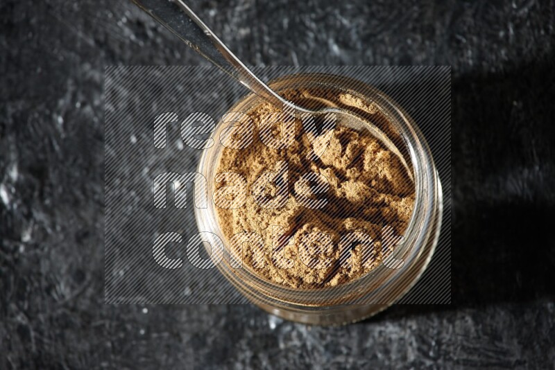 A glass jar and a metal spoon full of allspice powder on a textured black flooring