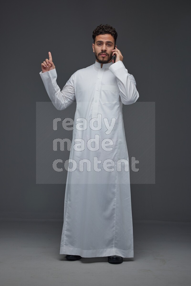 A Saudi man wearing Thobe and talking in the phone while standing and making different hand gestures eye level on a gray background