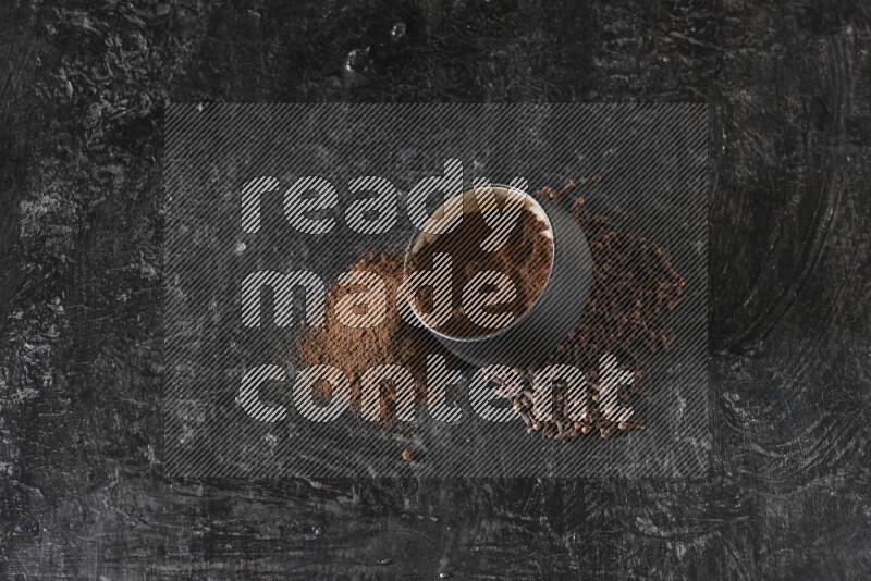 A black pottery bowl full of cloves powder spilled out of it with whole grains on a textured black flooring