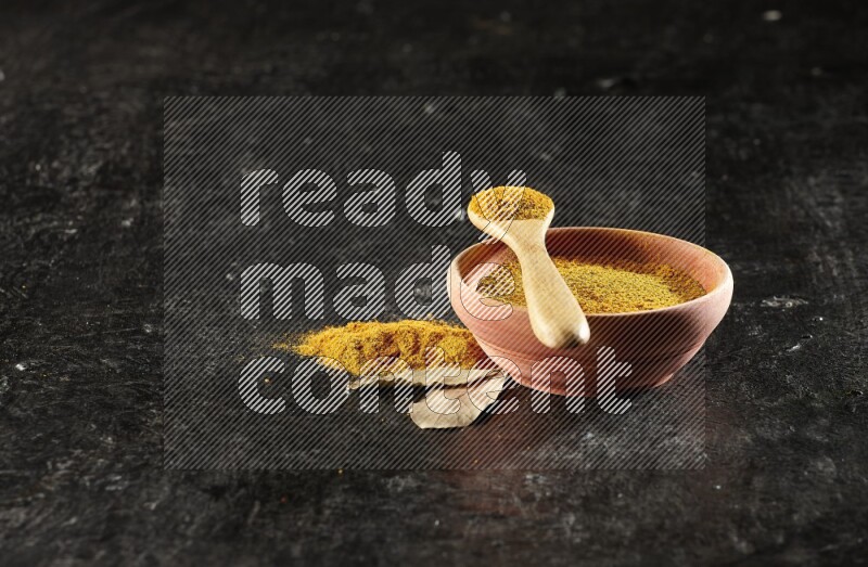 A wooden bowl and a wooden spoon full of turmeric powder on textured black flooring