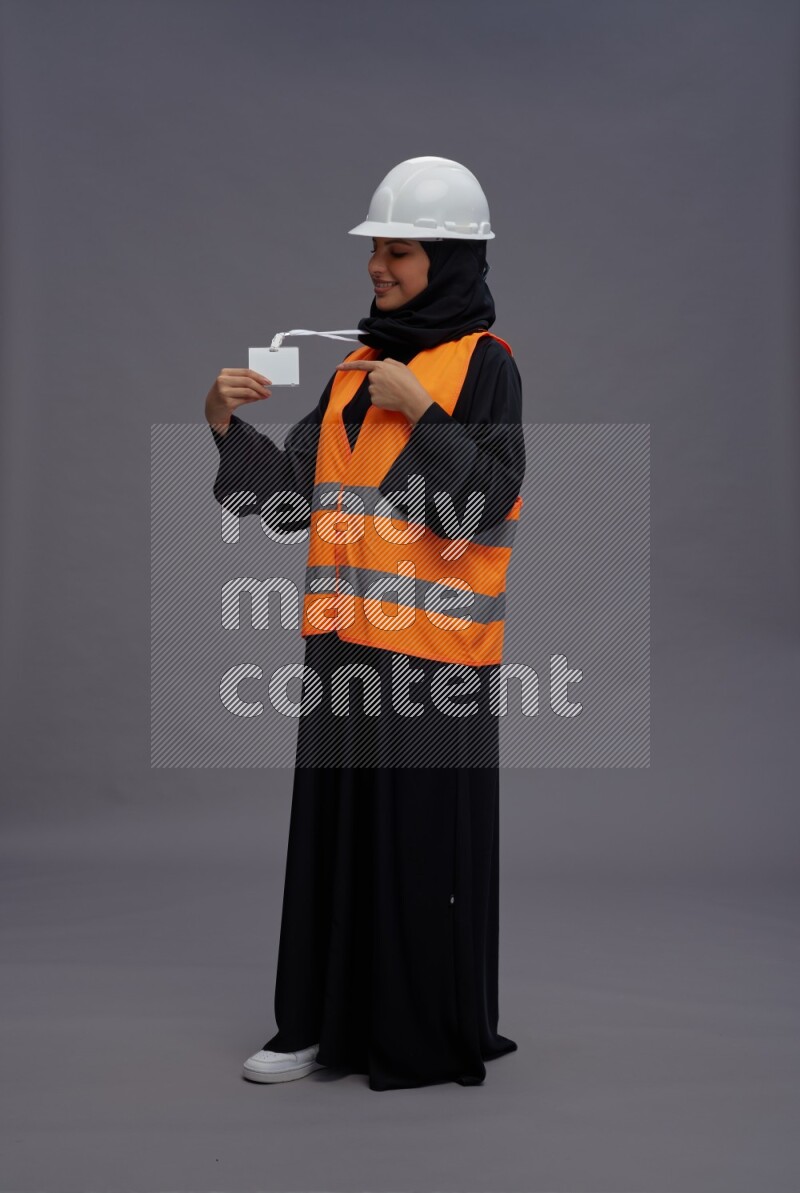 Saudi woman wearing Abaya with engineer vest with neck strap employee badge standing interacting with the camera on gray background