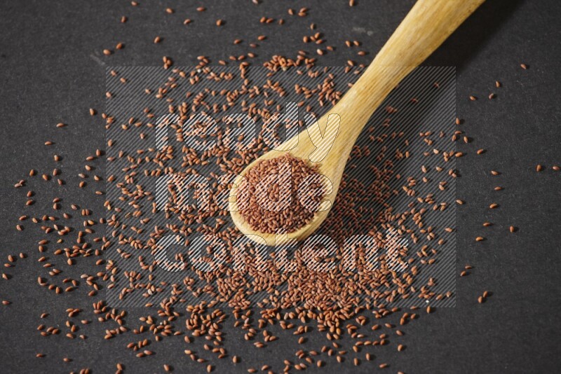 A wooden spoon full of garden cress seeds and seeds spread beside it on a black flooring
