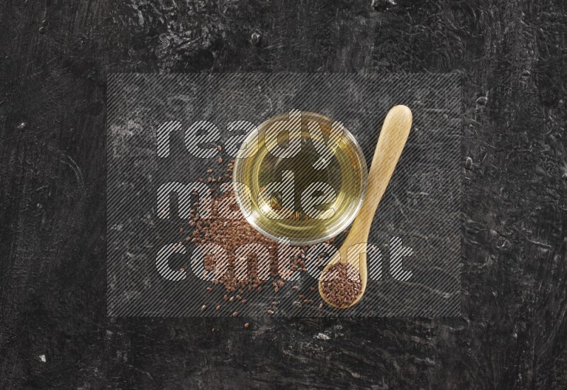 A glass bowl full of flaxseeds oil and wooden spoon full of flaxseeds with seeds spread on a textured black flooring