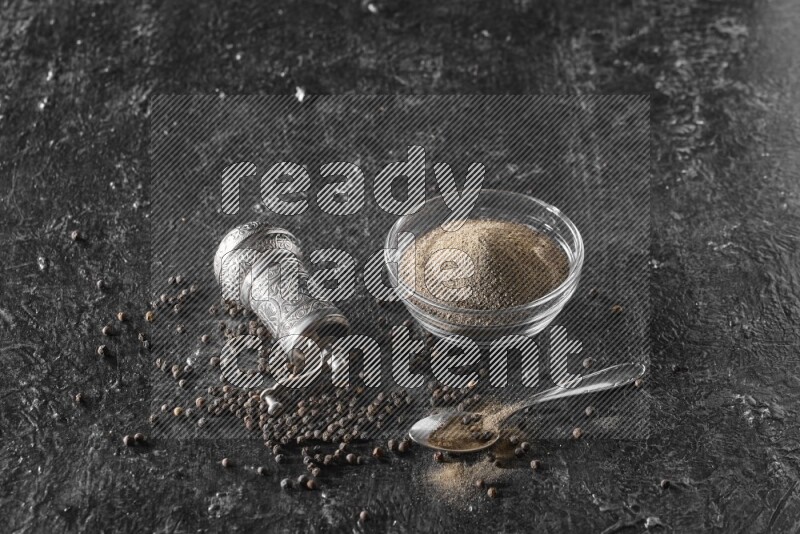 A glass bowl full of black pepper powder with black pepper beads, a turkish metal pepper grinder and a metal spoon on textured black flooring