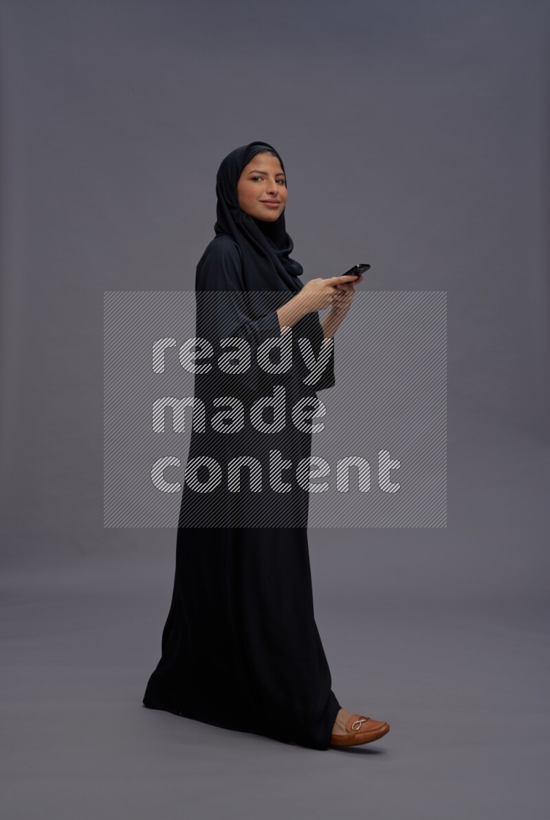 Saudi woman wearing Abaya standing texting on phone on gray background