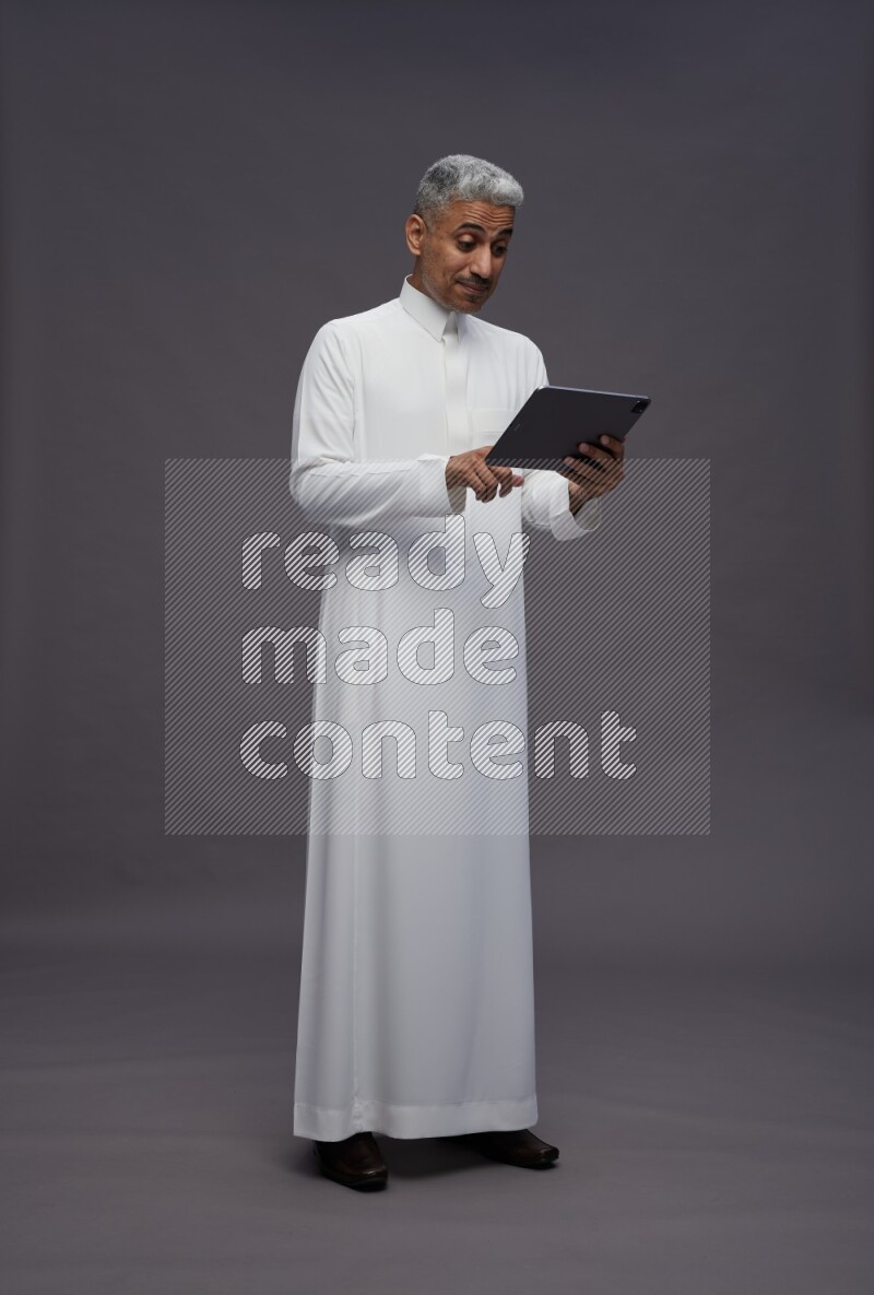 Saudi man wearing thob standing working on tablet on gray background