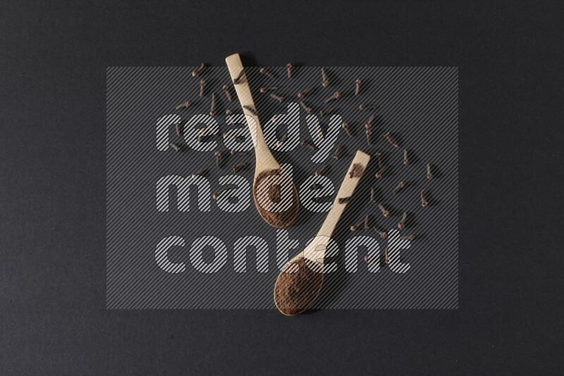2 wooden spoons full of cloves powder with spreaded whole cloves on a black flooring