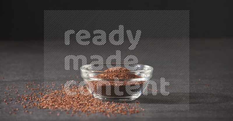 A glass bowl full of garden cress seeds surrounded by seeds on a black flooring