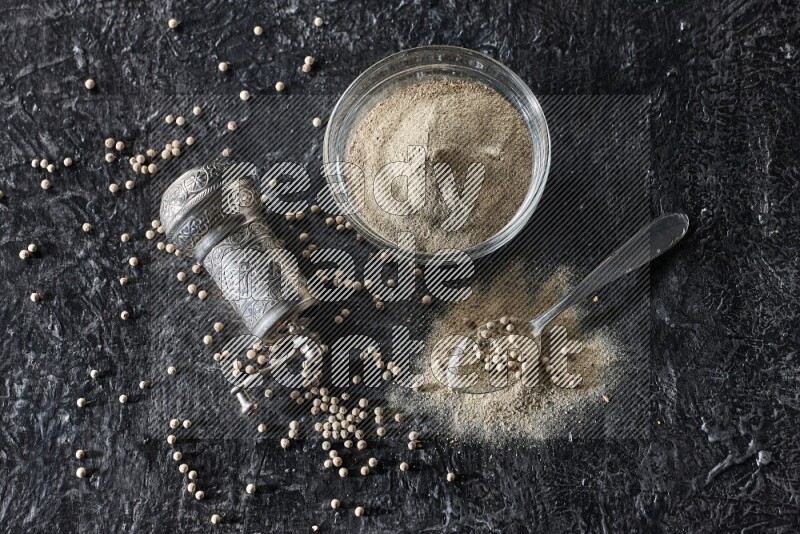 A glass bowl full of white pepper powder with pepper beads, a metal grinder and a metal spoon on textured black flooring