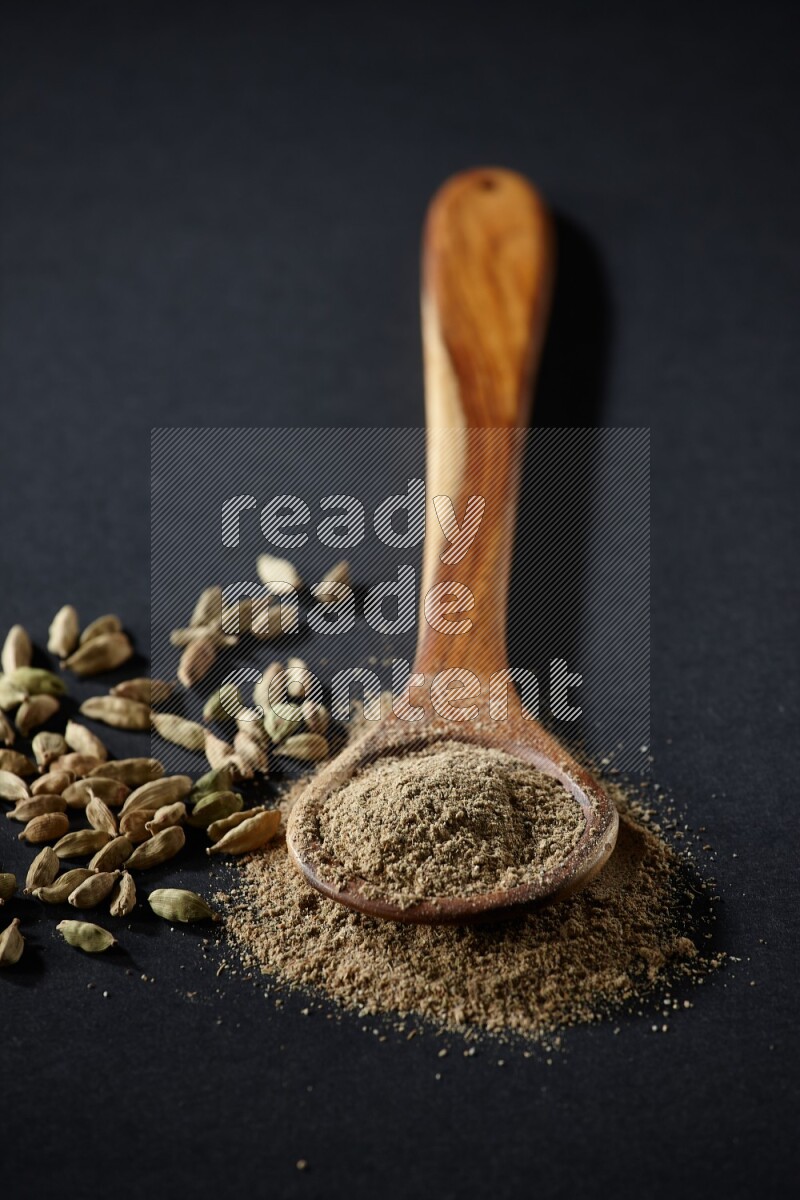 A wooden ladle full of cardamom powder and cardamom seeds beside it on black flooring