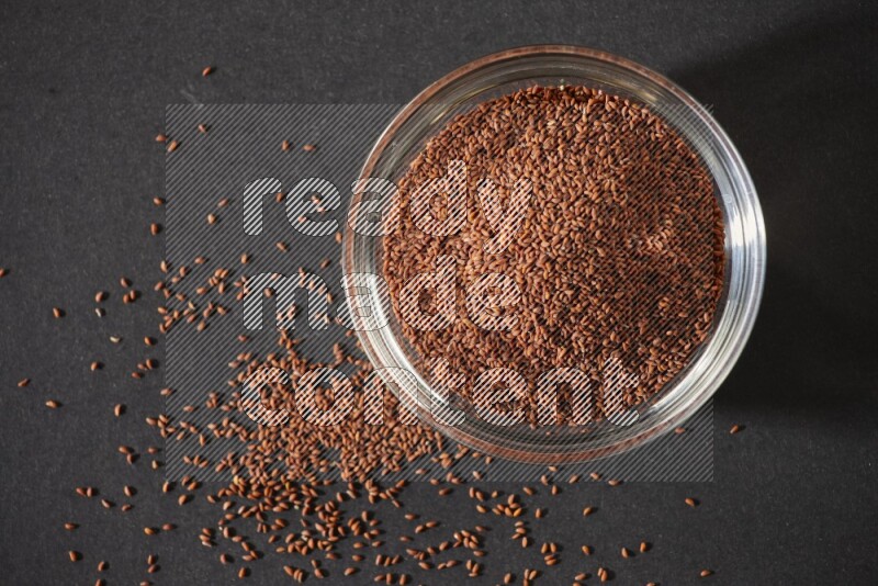 A glass bowl full of garden cress seeds surrounded by seeds on a black flooring