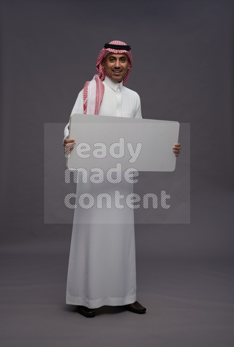 Saudi man wearing thob and shomag standing holding board on gray background
