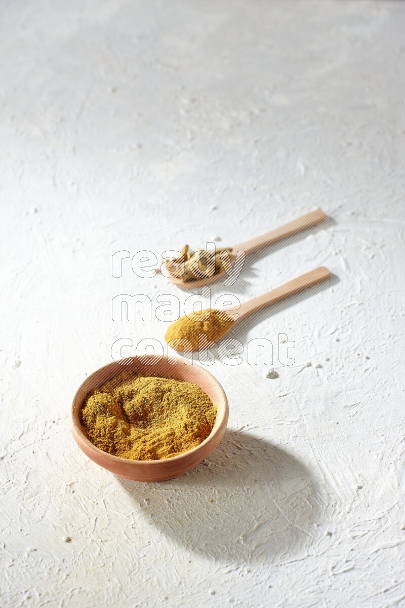 A wooden bowl full of turmeric powder and 2 wooden spoons full of dried turmeric whole finger and turmeric powder on textured white flooring