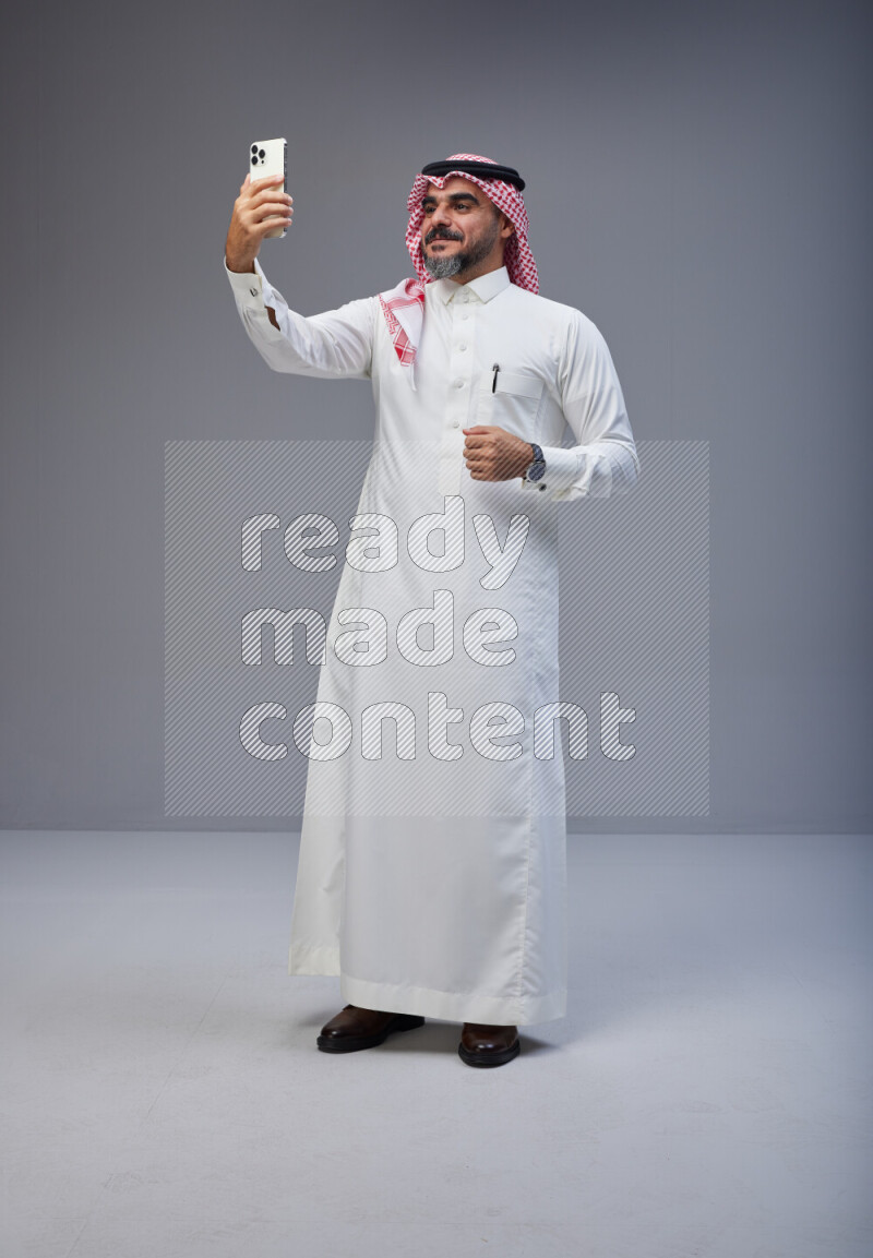 Saudi man Wearing Thob and red Shomag standing taking selfie on Gray background