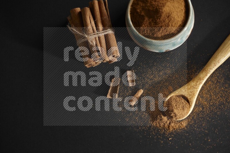 Ceramic bowl full of cinnamon powder and a wooden spoon full of powder with cinnamon sticks stacked and bounded on black background