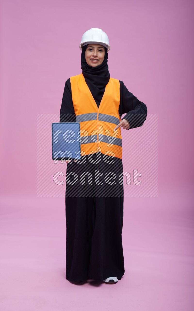 Saudi woman wearing Abaya with engineer vest and helmet standing showing tablet to camera on pink background