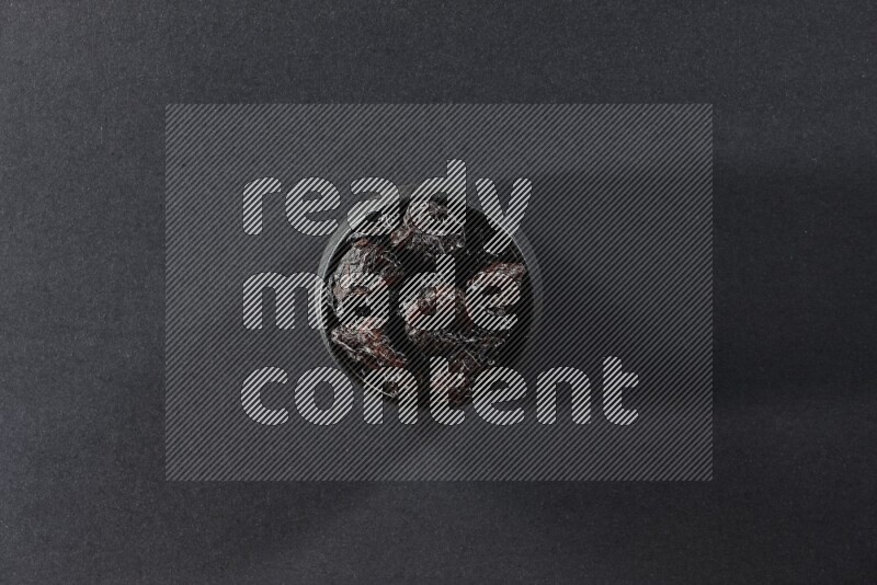 A black pottery bowl full of dried plums on a black background in different angles