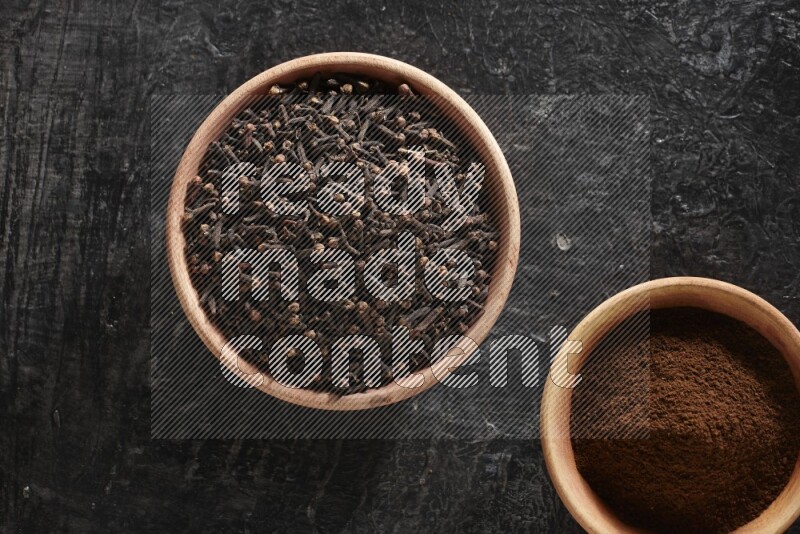 2 wooden bowls full of cloves powder and whole cloves on a textured black flooring