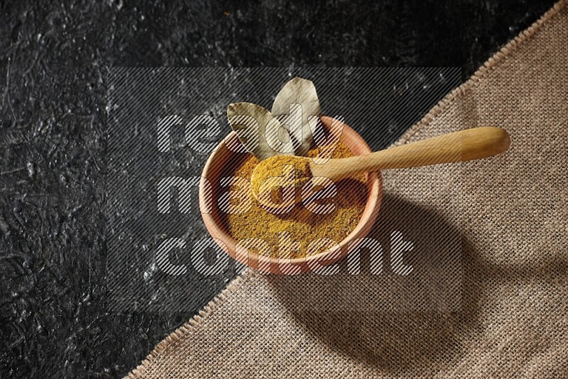 A wooden bowl and a wooden spoon full of turmeric powder on burlap fabric on textured black flooring