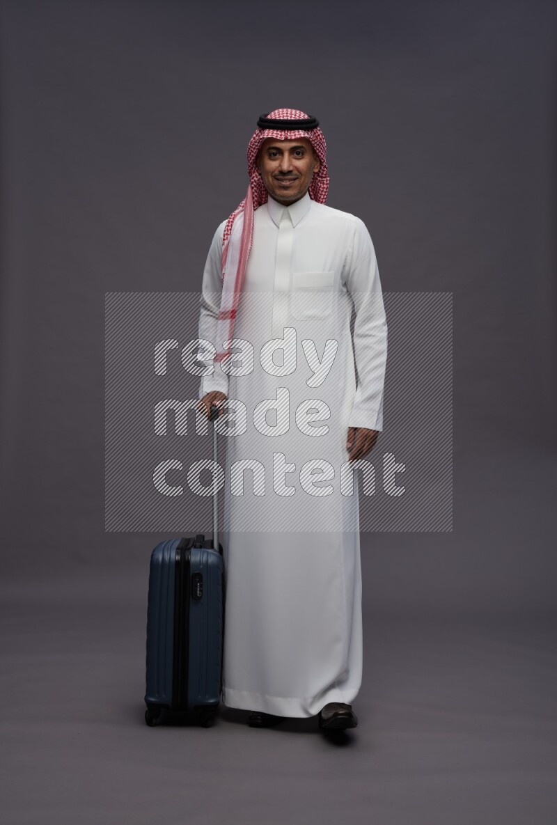 Saudi man wearing thob and shomag standing holding bag on gray background