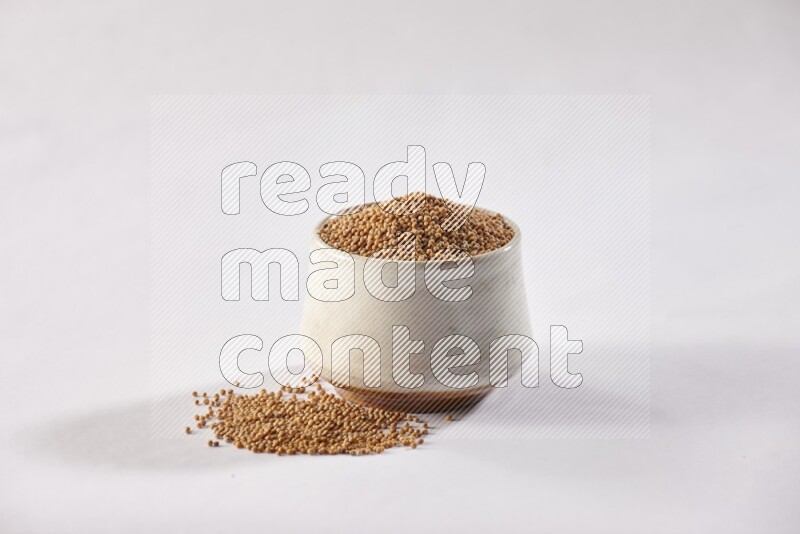 A beige pottery bowl full of mustard seeds and more seeds spread on a white flooring